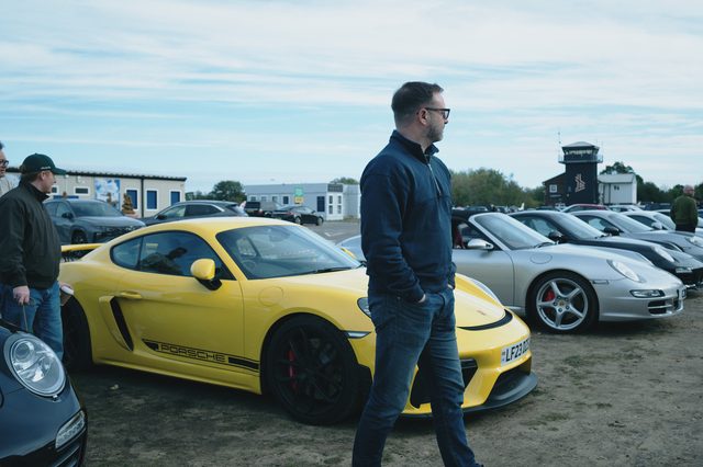 A man walks past a yellow Porsche on the dirt car park.
