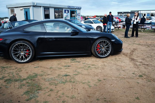A black Porsche 911 side on, people by the clubhouse behind.