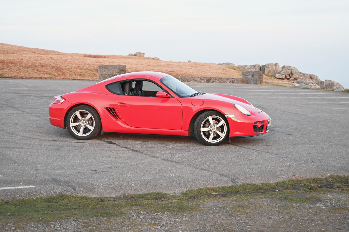 A red Cayman side on at the edge of a car park, sky behind.