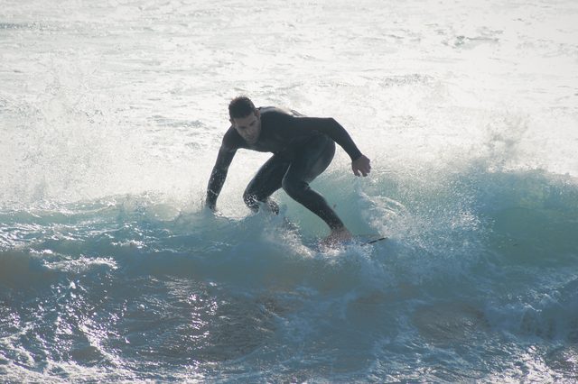 A skimboarder in a black wetsuit rides through white water, arms out for balance.