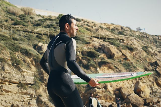 A skimboarder holds a green board in front of the cliffs, looking out to sea.