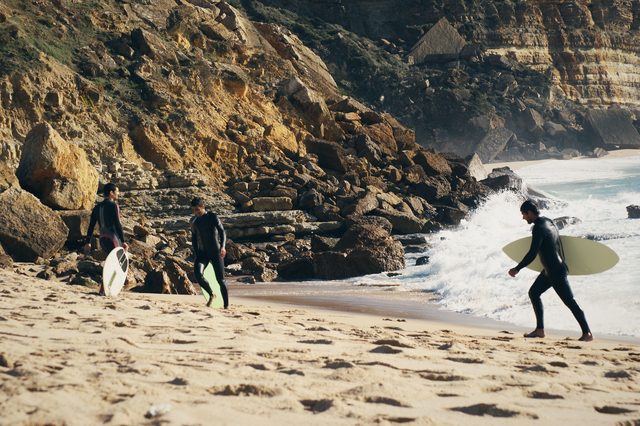 Three skimboarders on the sand at the foot of the rocks, a wave breaking behind them.