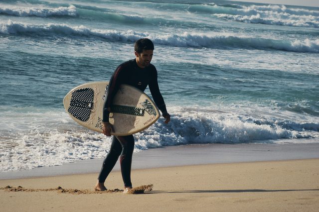 A skimboarder walks up the beach with his board under one arm.