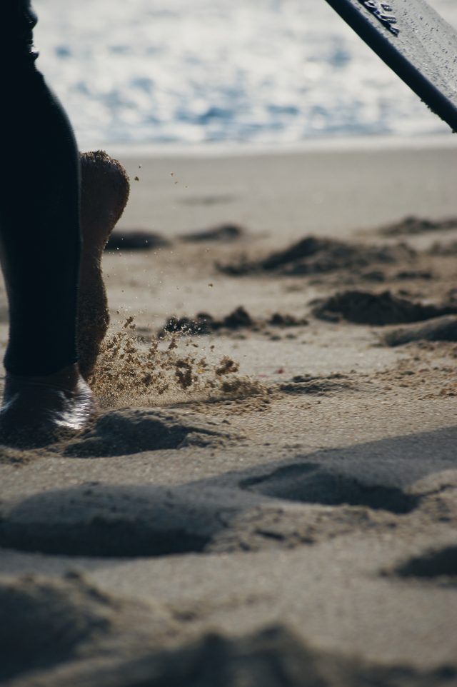 A bare foot kicks up sand, the tail of a board in the corner.