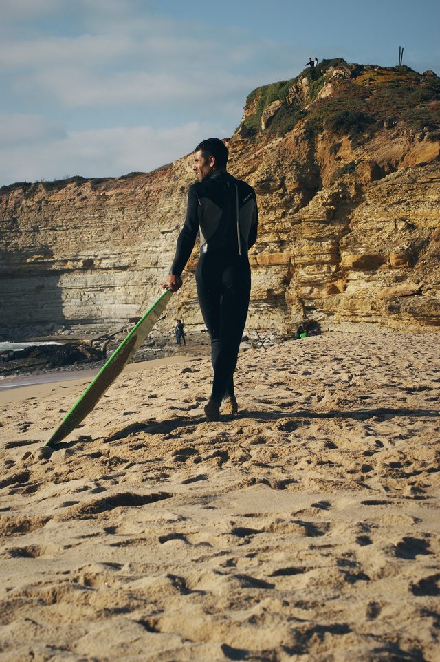 A skimboarder stands on the sand below the cliffs, board at his side.