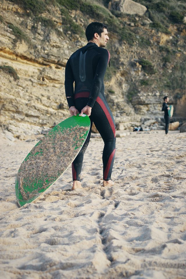 A skimboarder holds a green speckled board, another figure on the beach behind.