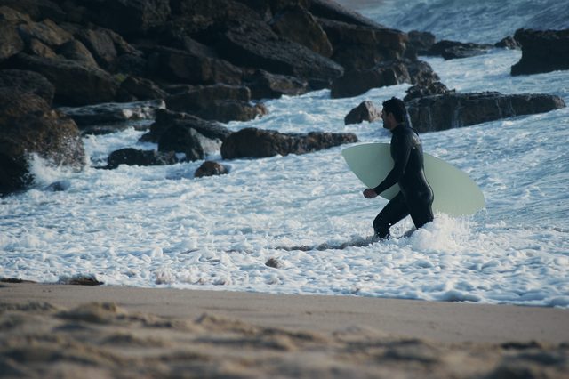 A man wades out among the rocks and white water carrying a board.