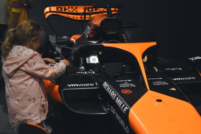 A young girl in a pink jacket reaches into the cockpit of a papaya racing car.