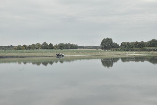 A still lake under a grey sky, trees on the far bank reflected in the water, a small car parked at the water's edge.