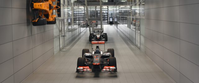 A long white corridor of racing cars: one car on the floor in front, another behind it, a papaya car mounted high on the wall.