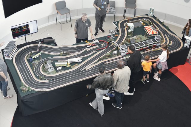 Seen from above, families gather round a big slot-car track; a man races while two children watch from the rail.