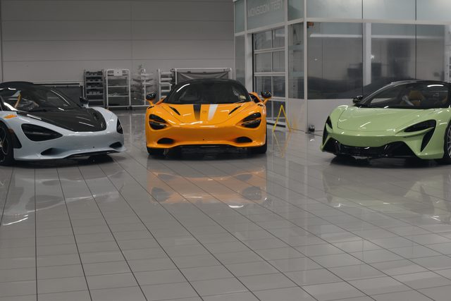 Three finished sports cars, pale blue, papaya and green, waiting in a row on a white tiled floor.