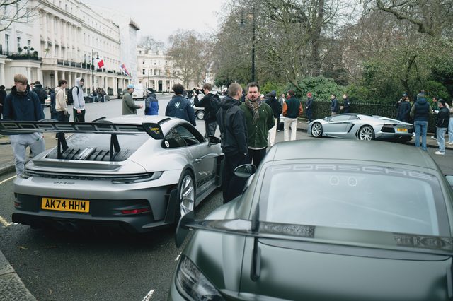 A crowd gathers around sports cars on a London square lined with white stucco houses.