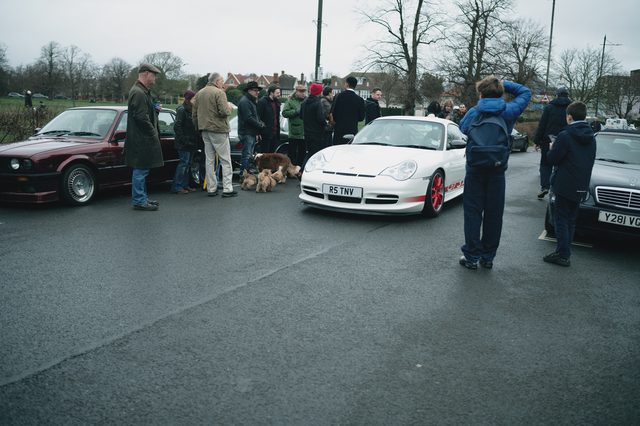 People and their dogs stand around a white Porsche on a grey morning.