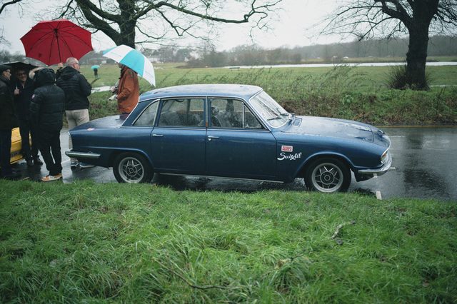 People under red and white umbrellas around a dark blue saloon parked on wet grass.
