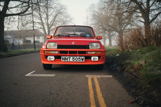 A red hatchback faces the camera on double yellow lines in a misty, tree-lined road.