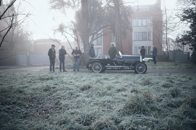 Owners stand around a vintage open racing car on frosted grass in the morning fog.