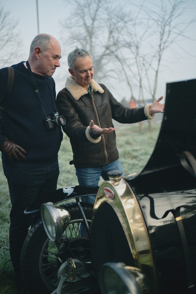 Two men in winter coats lean over a vintage car; one opens his hands to explain the engine.