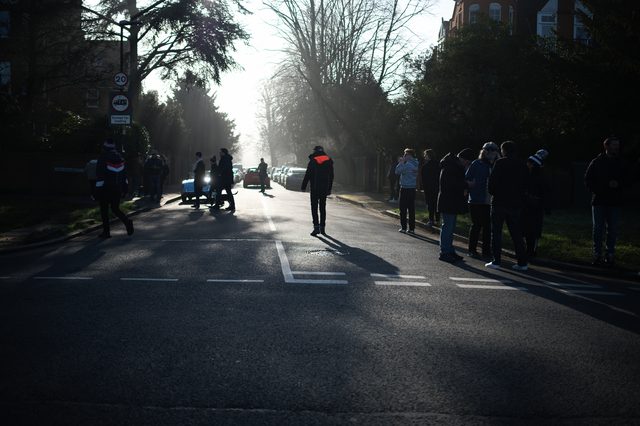 People and cars line a road that runs into hazy low sun.