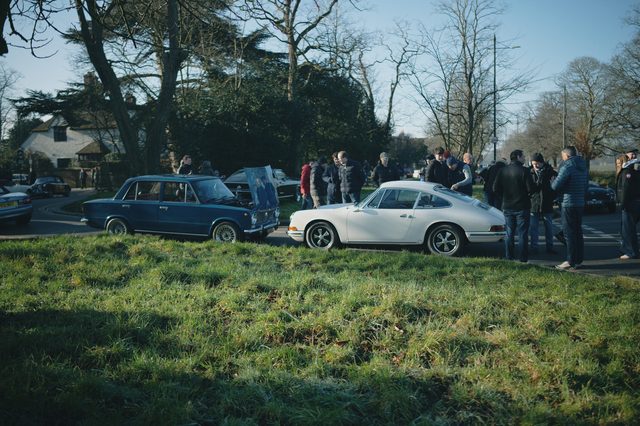 Owners gather around classic cars parked along the edge of a green.