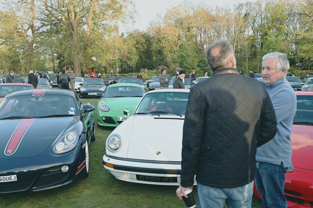 Two men talk among Porsches parked on the grass at an evening meet.