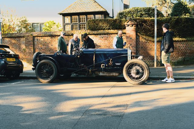 Owners talk around a vintage open tourer parked in morning sun.