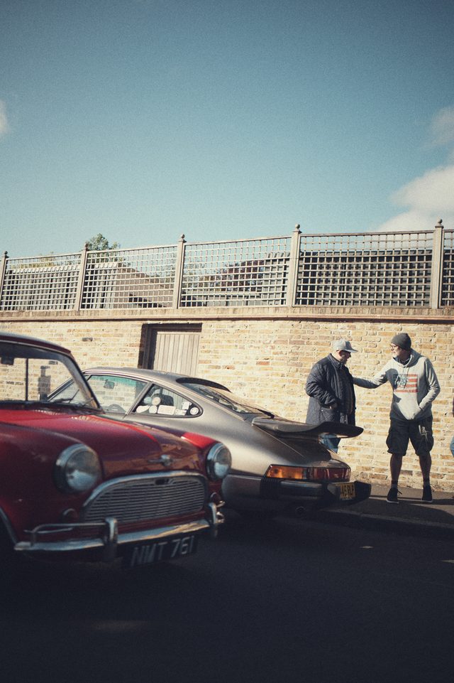 Two men talk beside a classic Porsche, a red classic car in front, under a pale sky.