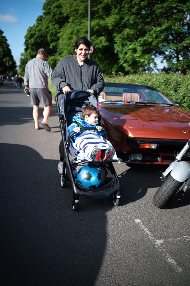 A man pushes a baby in a pram past a red sports car.