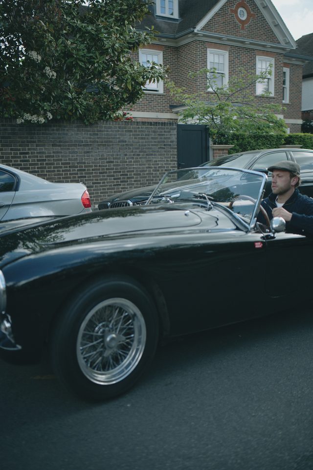 A man in a flat cap sits in a black open sports car.