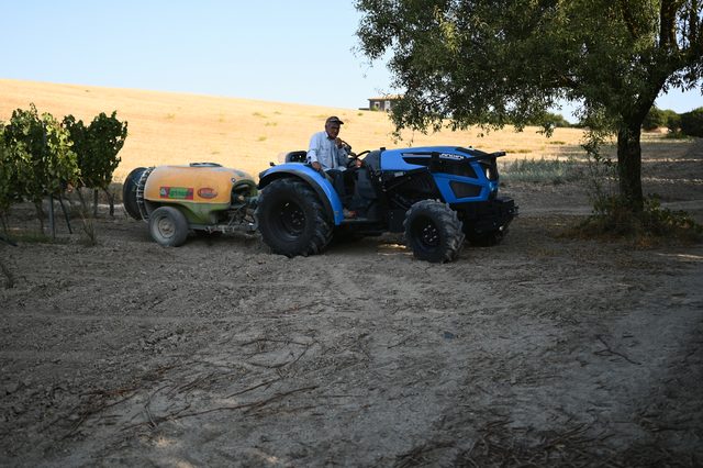 A man drives a blue tractor and sprayer along the edge of the vineyard.