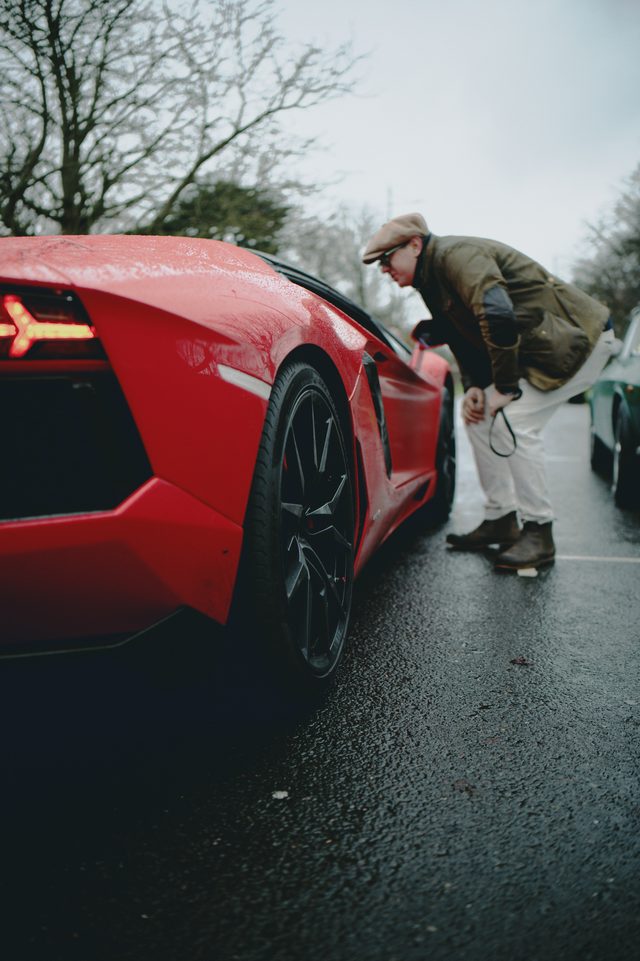 A man bends to look closely at the side of a red supercar on a wet road.