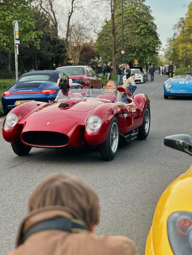Over a photographer's shoulder, a red vintage racing car drives down a road lined with people.